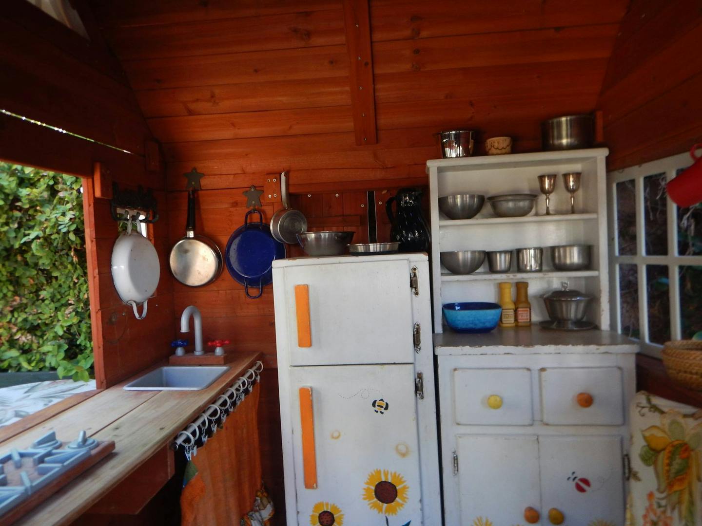 Play kitchen with wooden walls, sink, refrigerator, and shelves with shelves of pots and pans