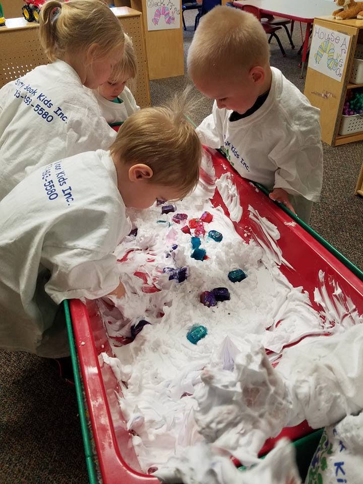 Three children playing with shaving cream and colorful objects in a red bin