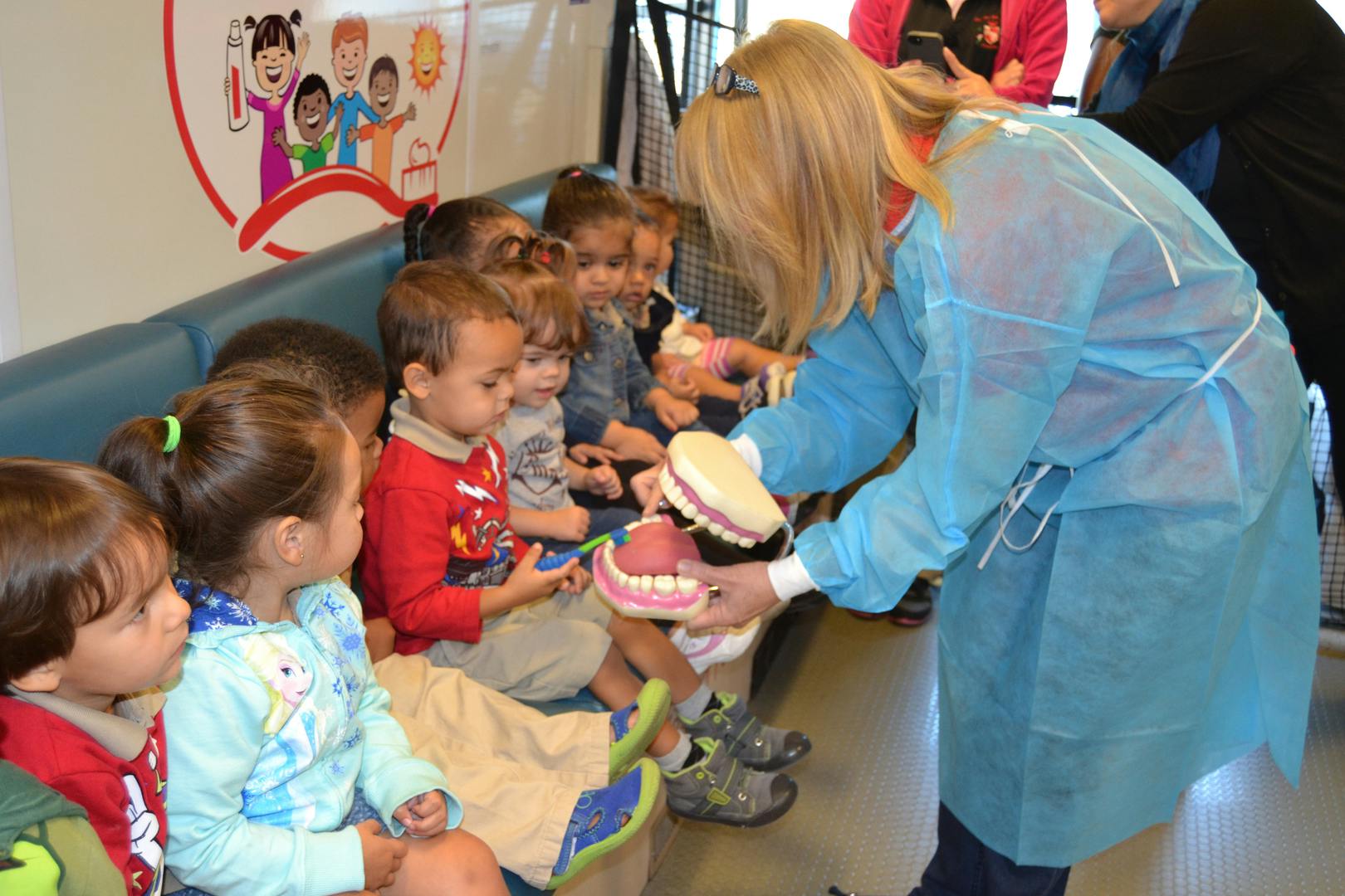 Person in scrubs demonstrating teeth brushing to a group of children
