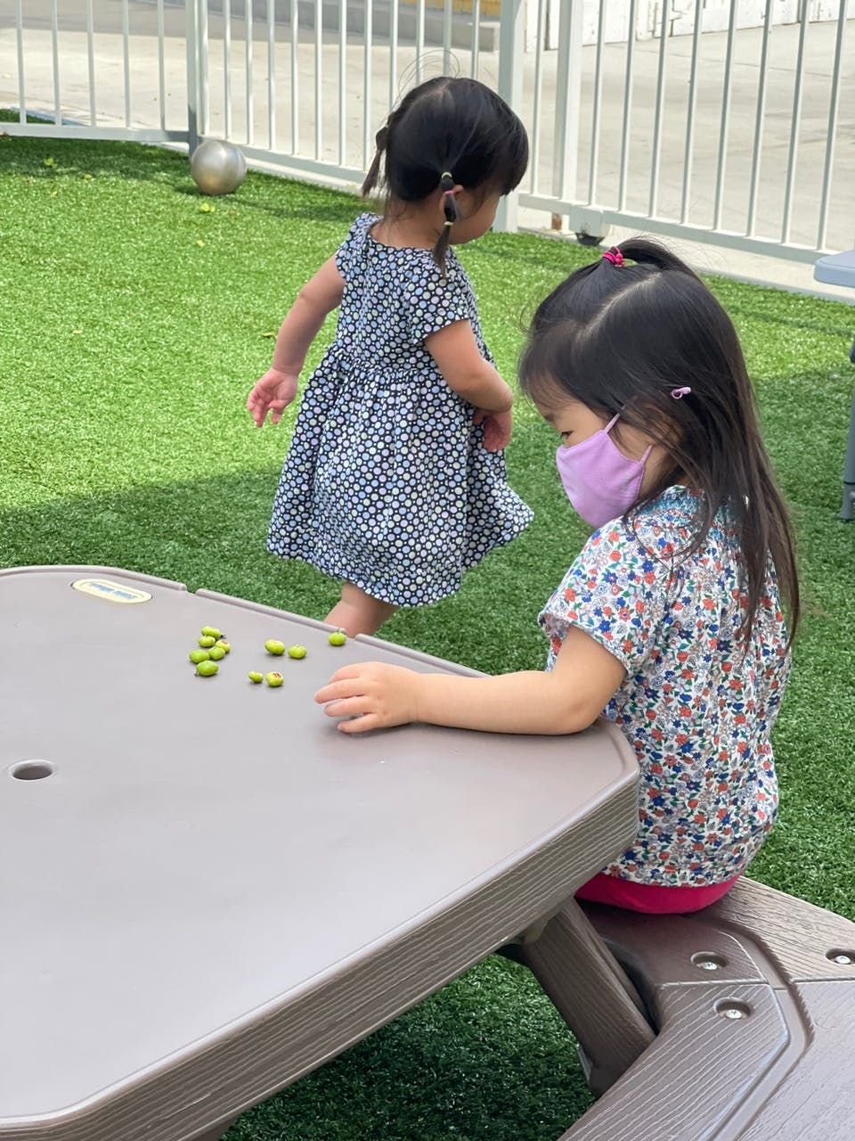 Child in mask sitting at table with another child walking nearby