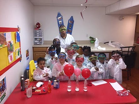 Adult and children in lab coats and safety goggles with balloons on a table