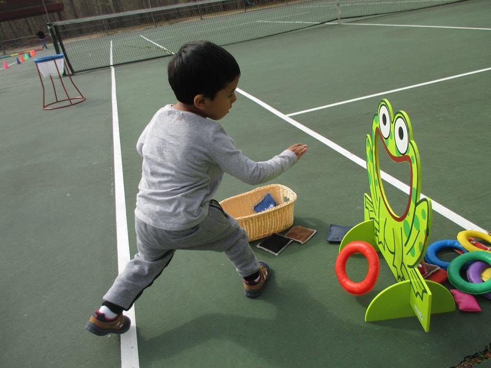 Child playing a ring toss game with a frog-shaped target on a tennis court