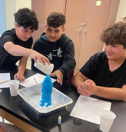 Three teens watching a volcano science experiment erupt in a classroom