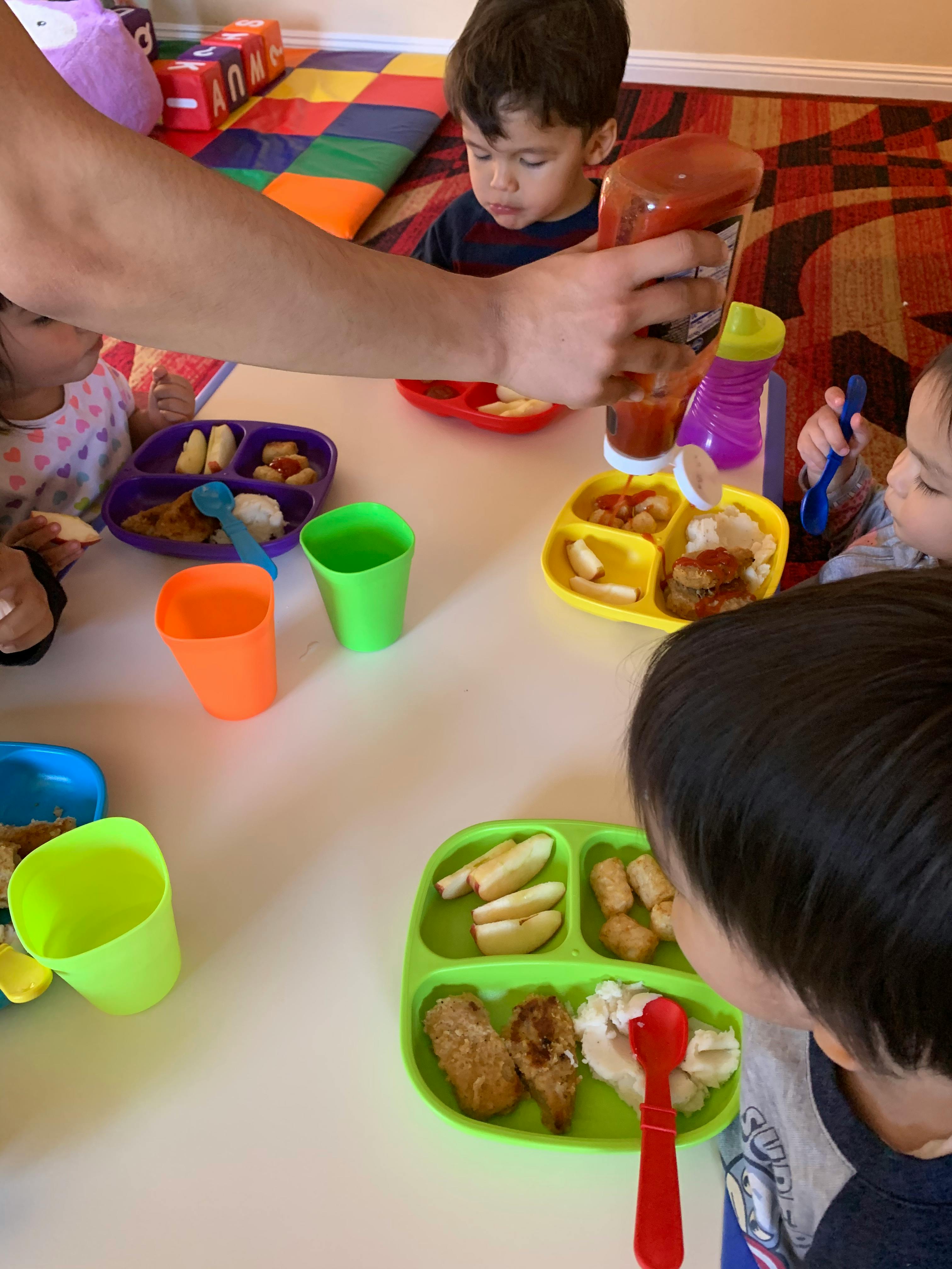 Person squeezing ketchup onto a child's plate at a table with other children eating