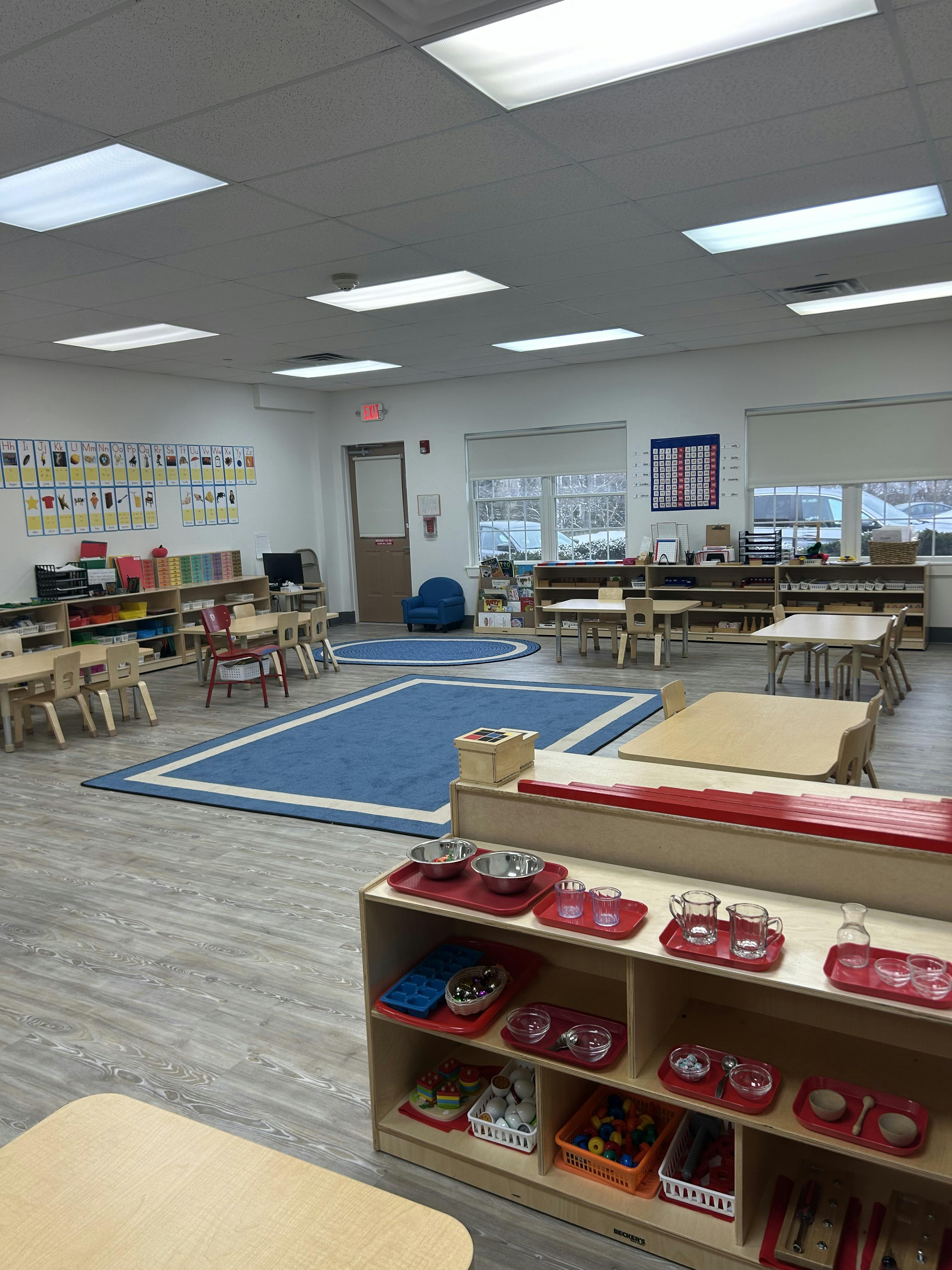 Classroom with small tables and chairs, shelves with learning materials, and a blue rug