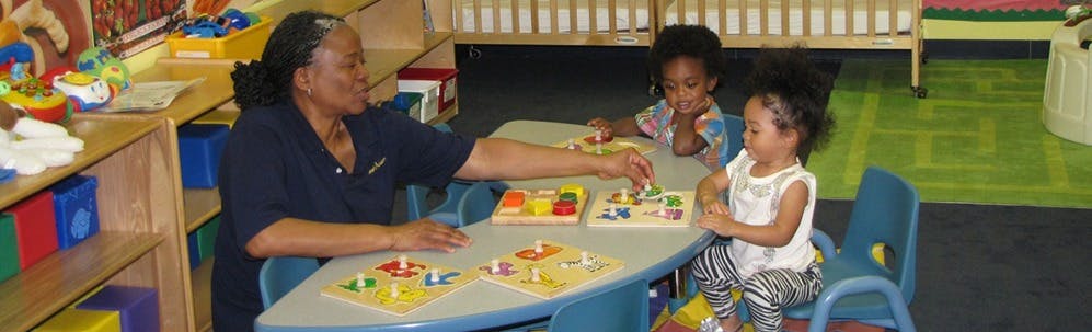 Person and two children playing with puzzles at a table in a classroom