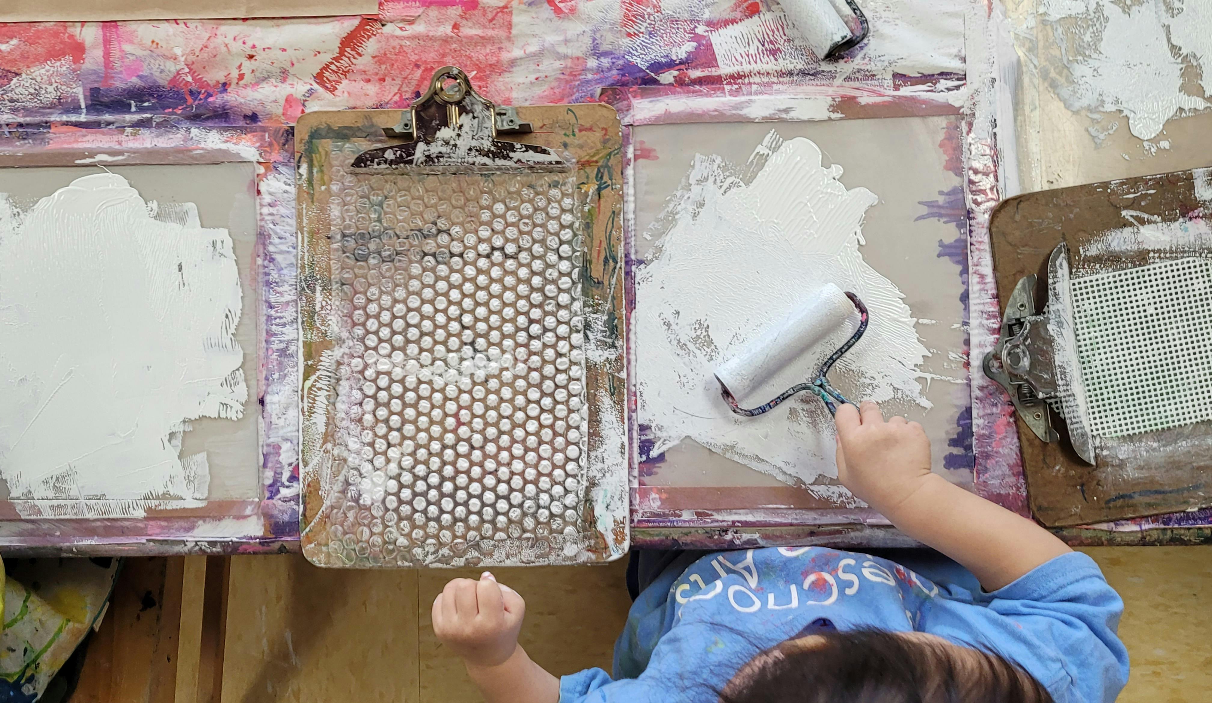 Child using a roller to apply white paint to a screen printing frame