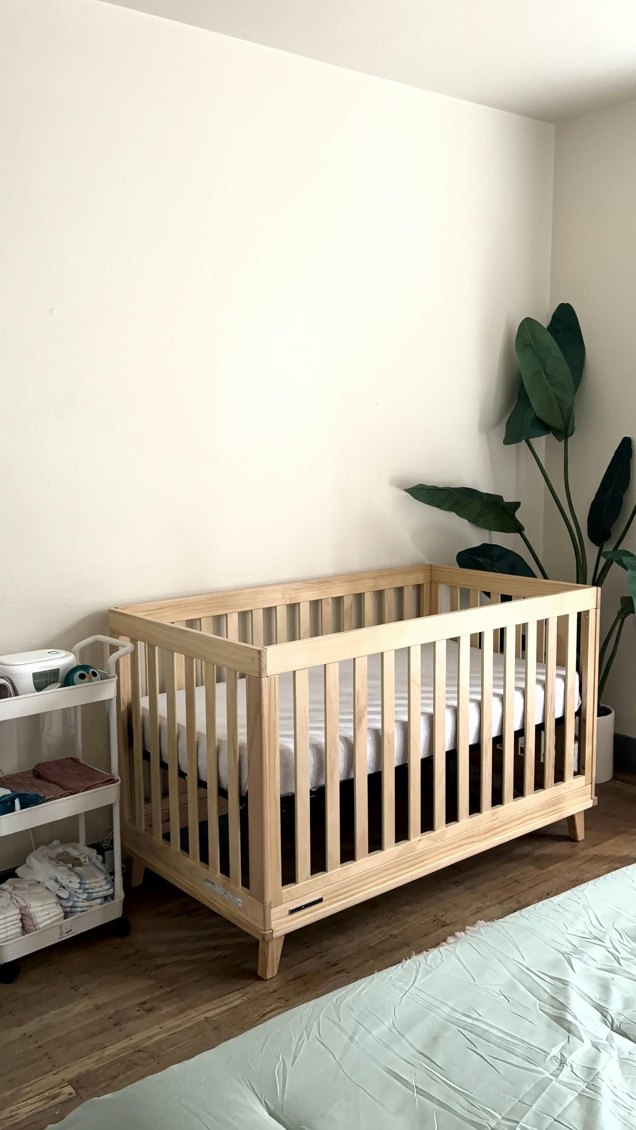 Wooden crib with a mattress next to a rolling cart with baby supplies