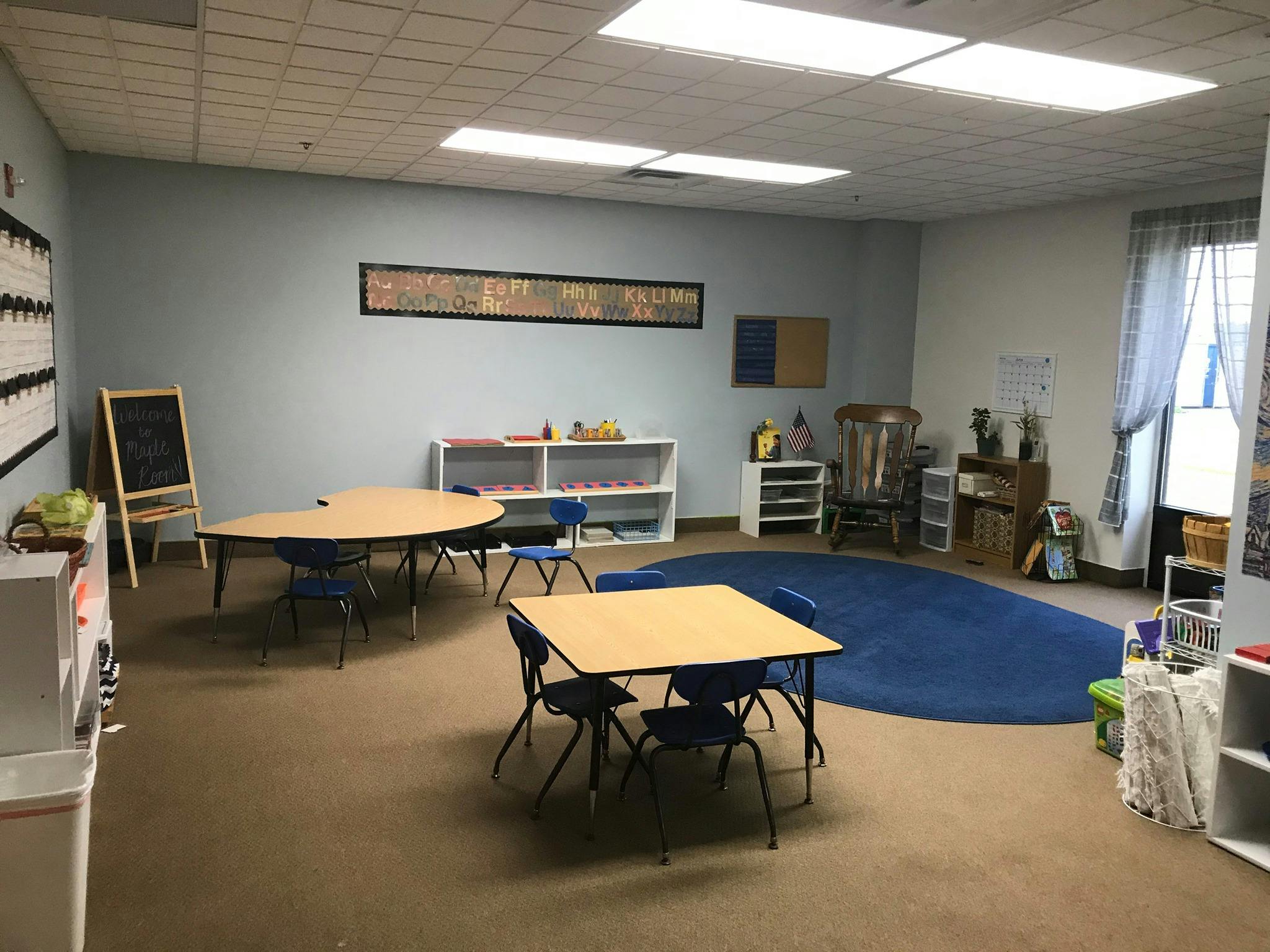Empty classroom with small tables and chairs, shelves, and a blue rug