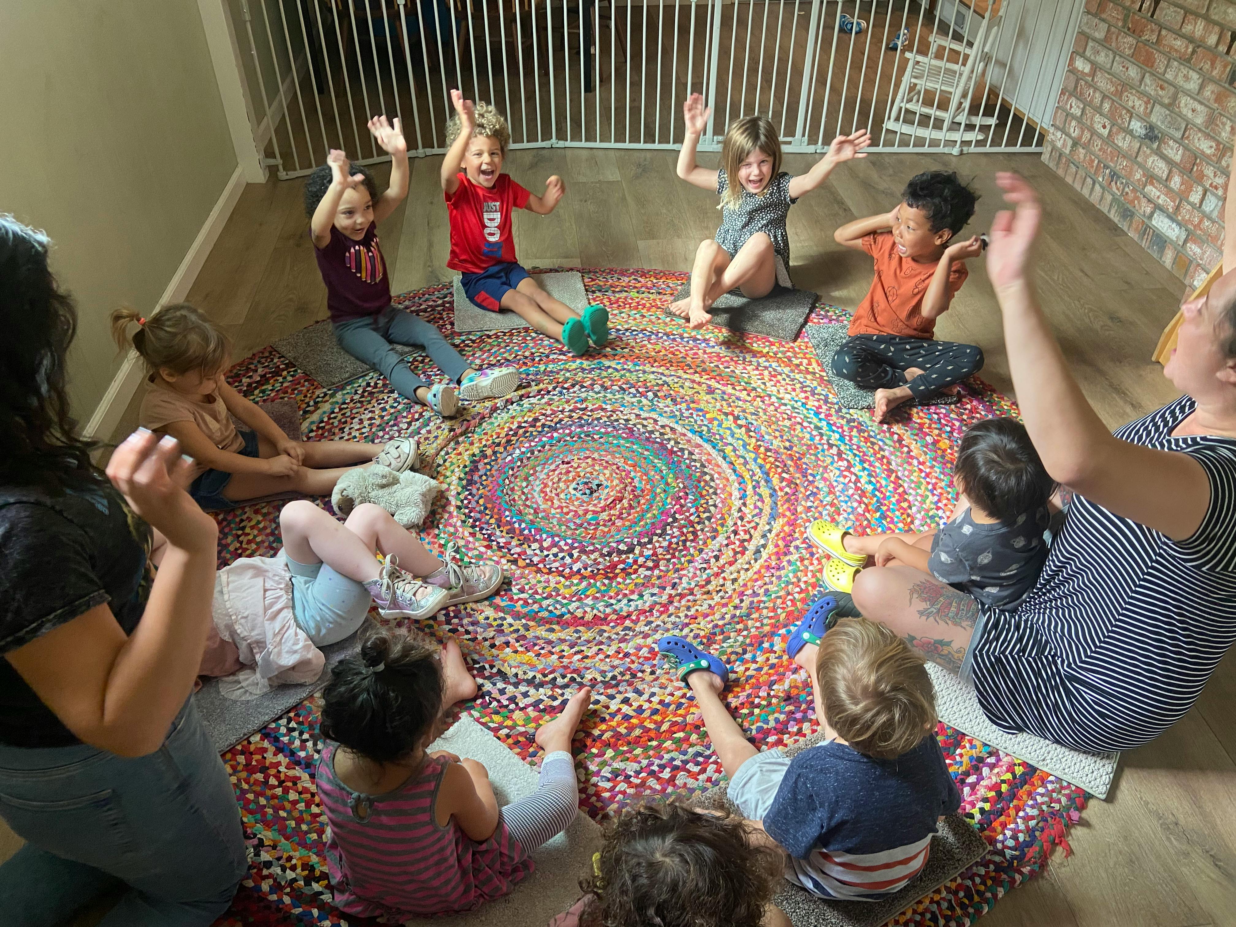 Adults and children sitting on colorful rugs with some raising their hands
