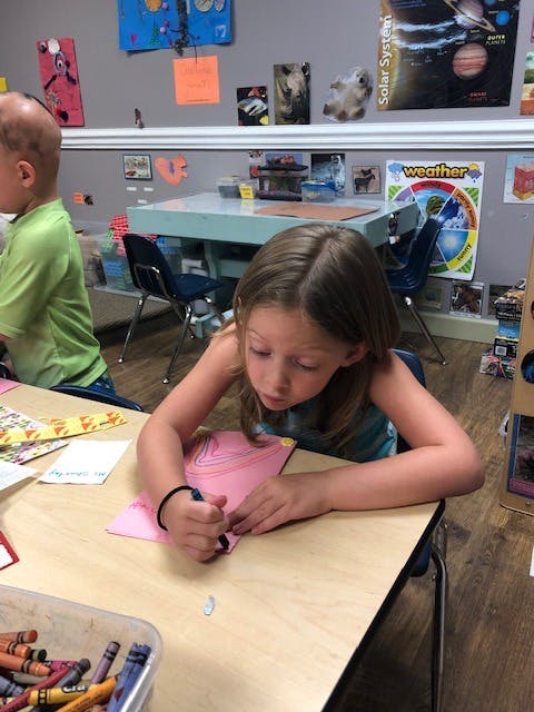 Child drawing on pink paper at a desk in a classroom