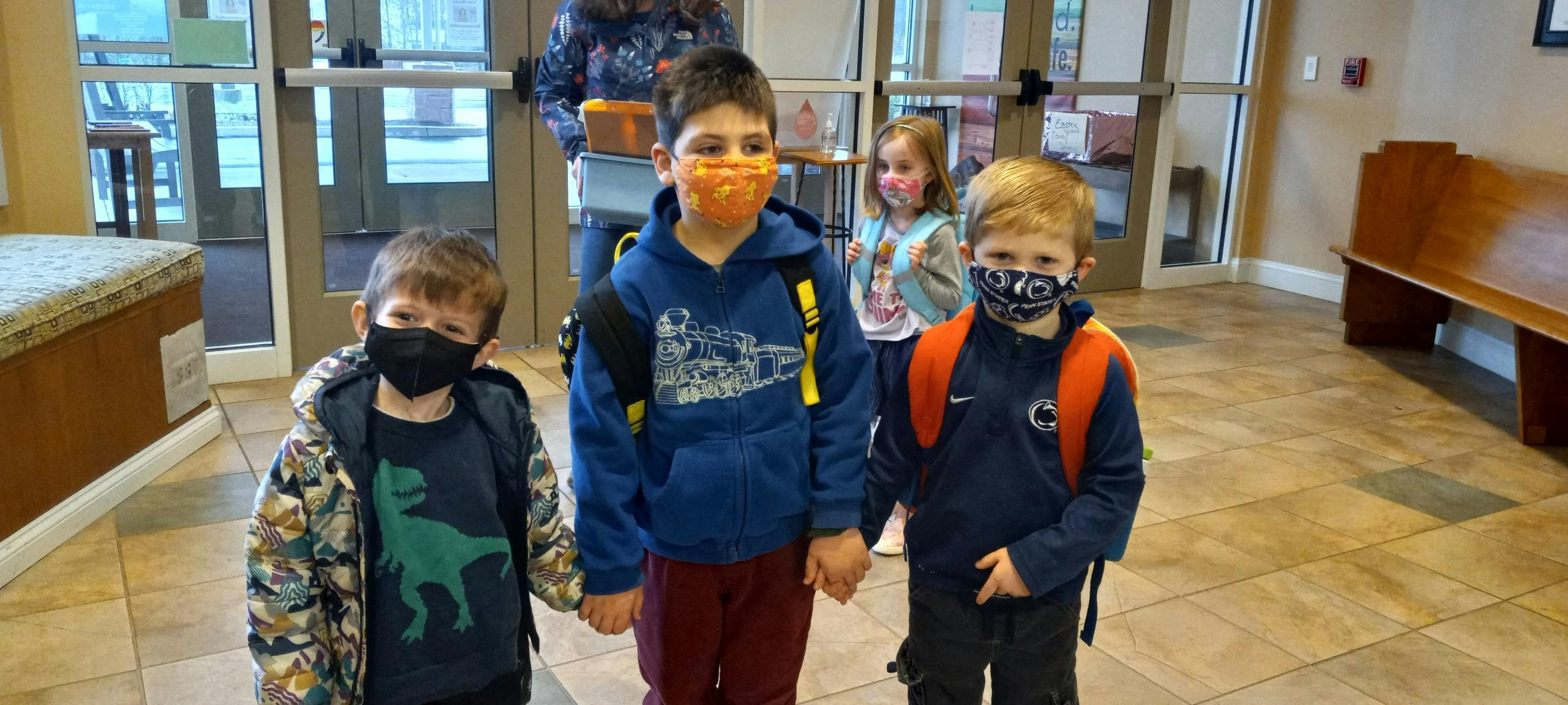 Three children wearing masks and backpacks standing indoors