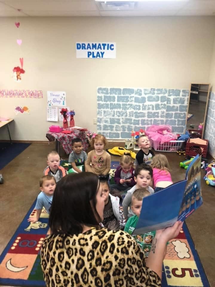Person reading a book to a group of children sitting on a rug in a classroom