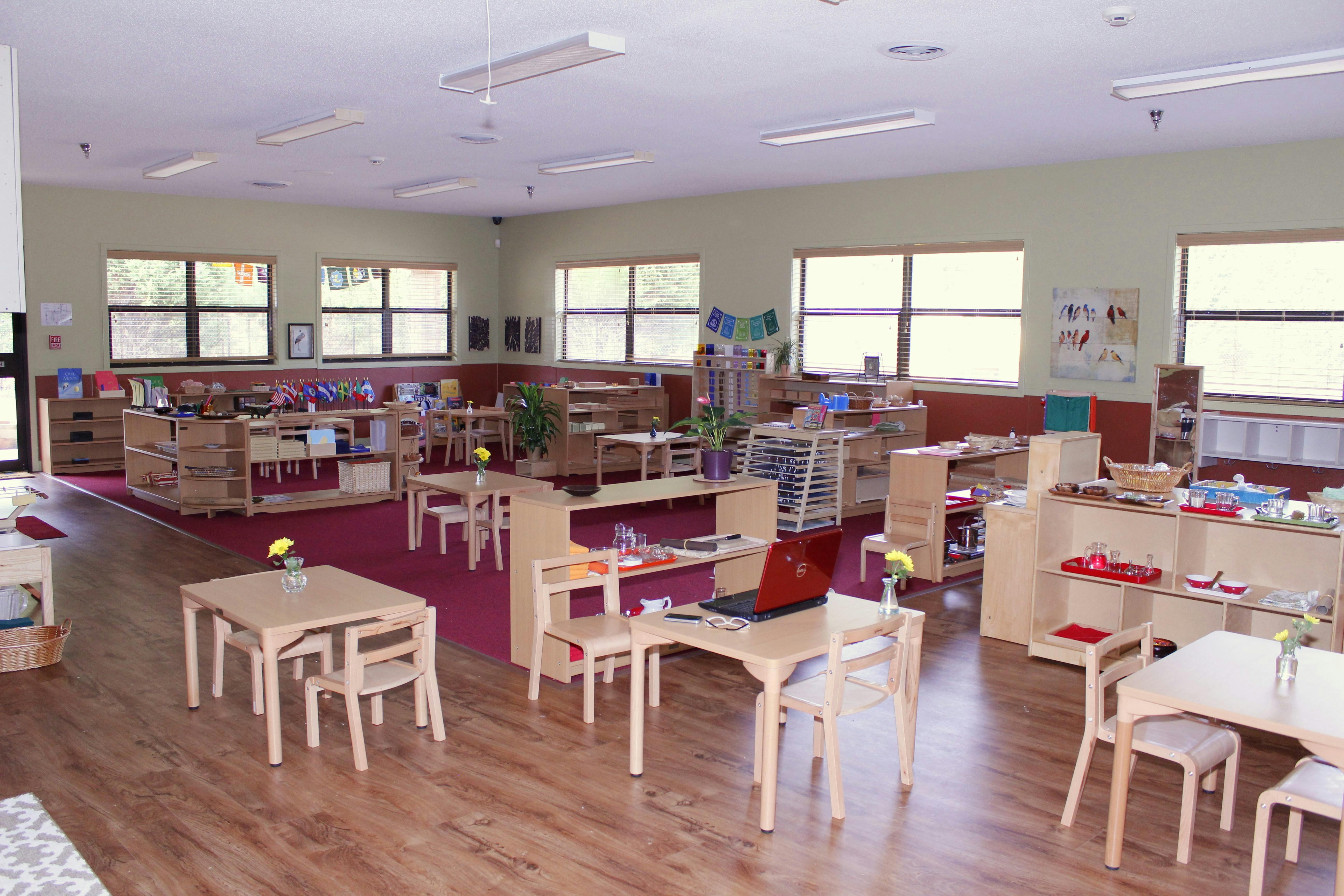 Classroom with small wooden tables and chairs, shelves with learning materials