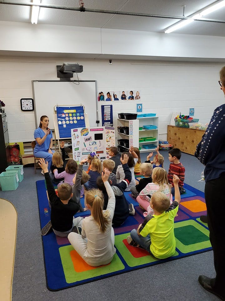 Person speaking to children sitting on a rug with hands raised in a classroom