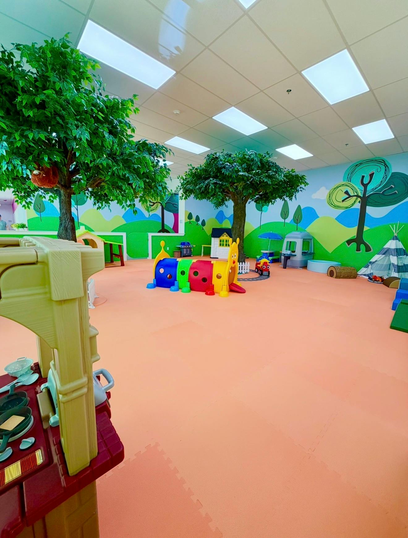 Indoor playground area with toy houses, a play tunnel, and artificial trees