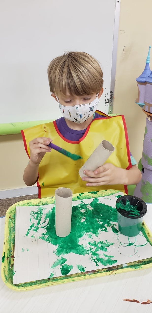 Child in a mask painting a cardboard tube with green paint