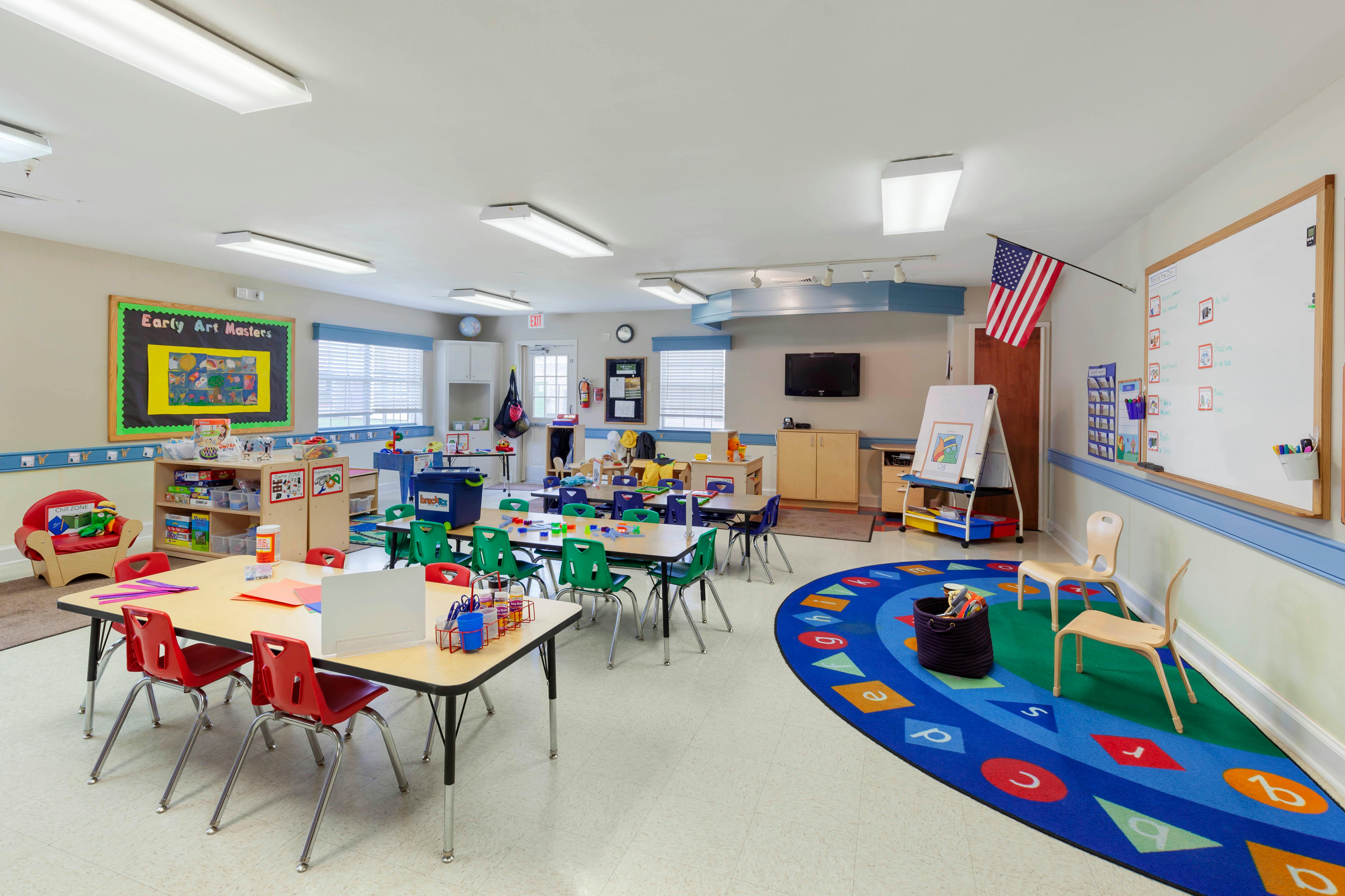 Colorful classroom with tables and chairs, a play rug, and an American flag