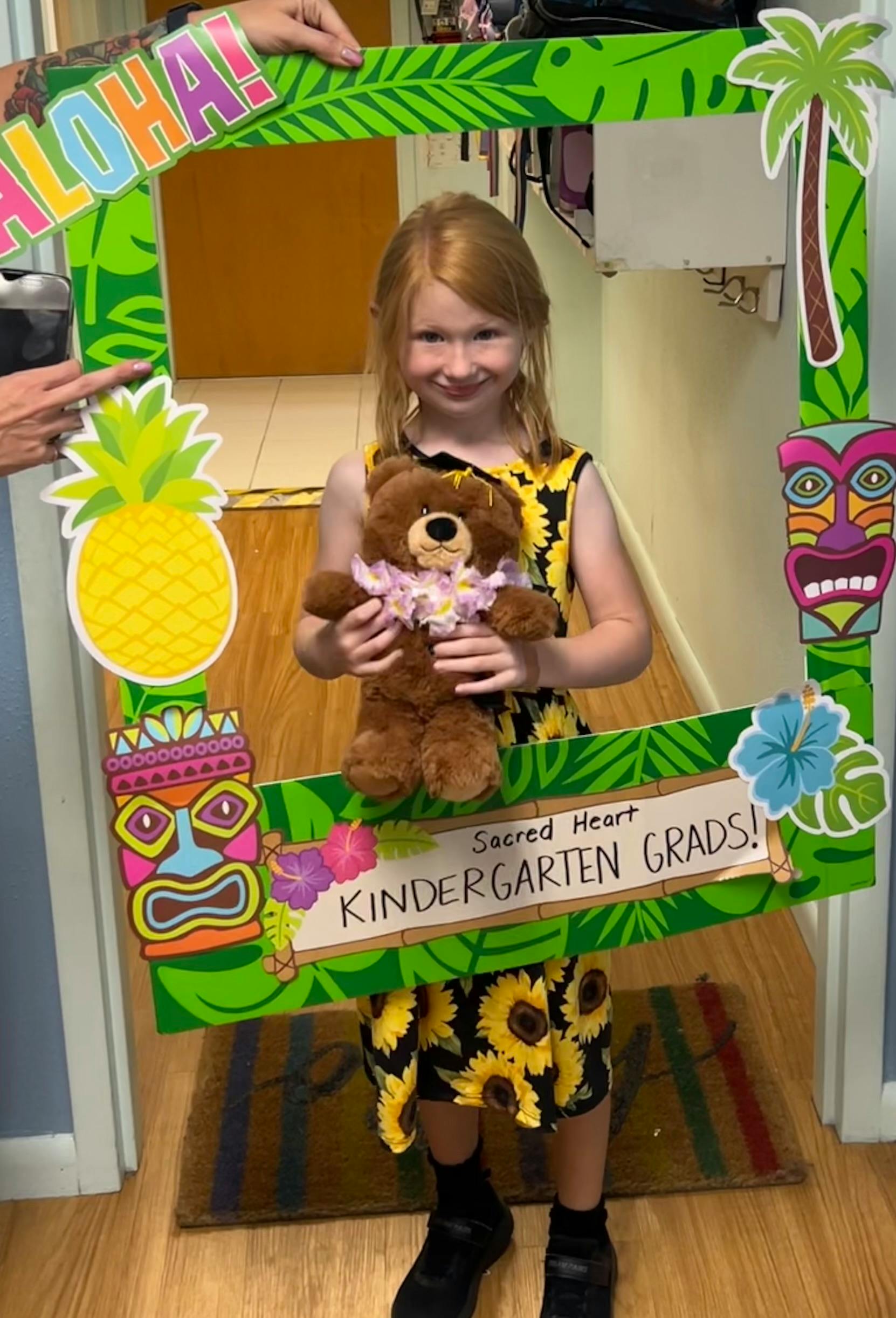 Child holding a teddy bear in a photo frame with "Kindergarten Grads" text