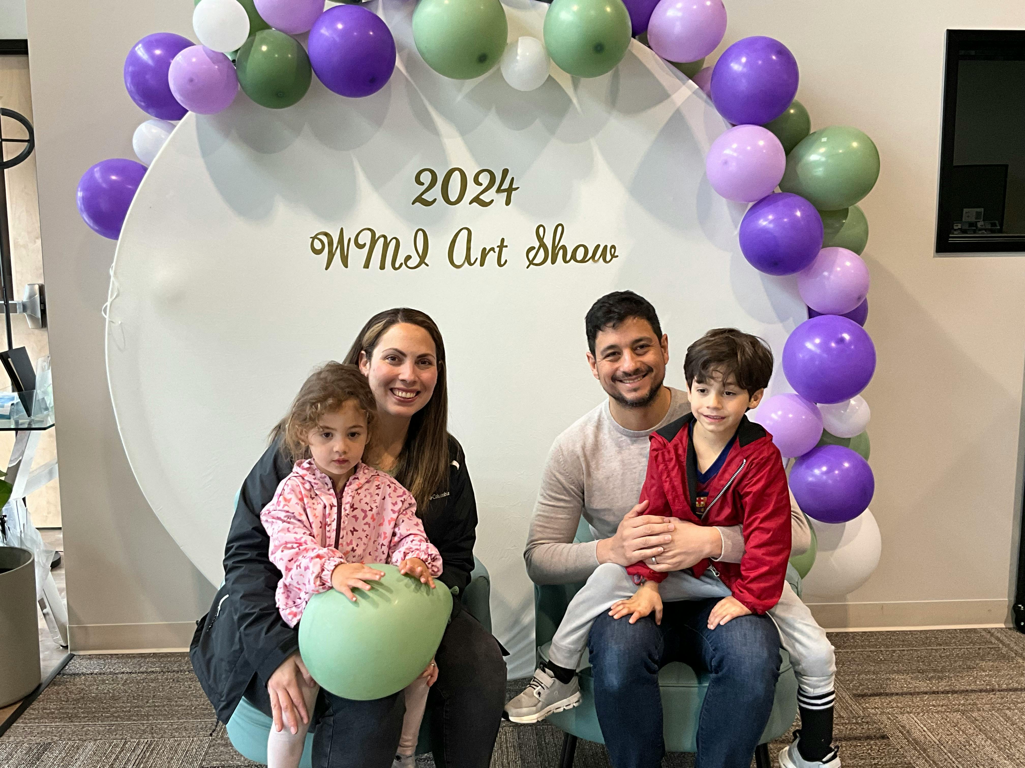 Two adults and two children sitting in front of a sign that reads "2024 WMI Art Show"