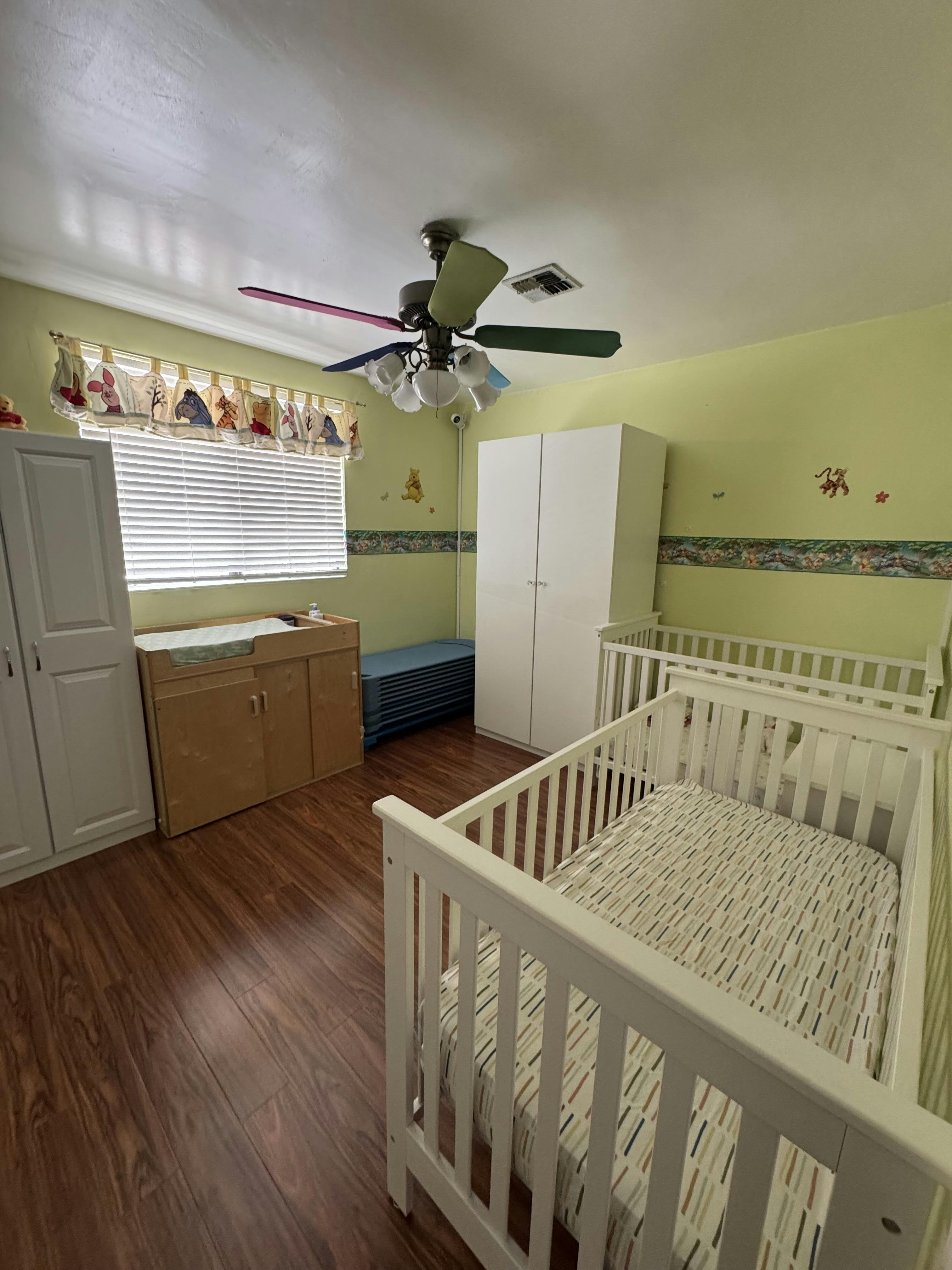 A room with two cribs, a changing table, and a colorful ceiling fan