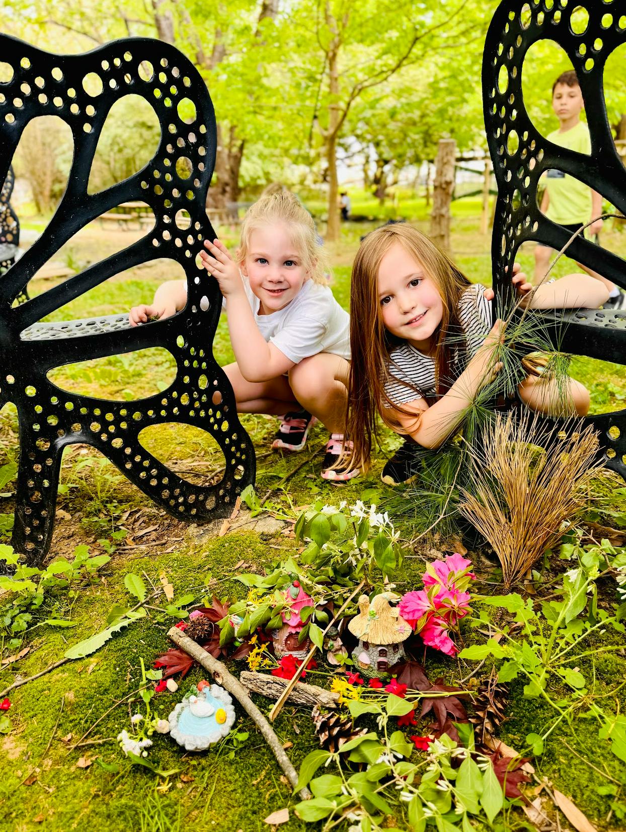 Two children crouching behind a butterfly-shaped bench with miniature fairy houses