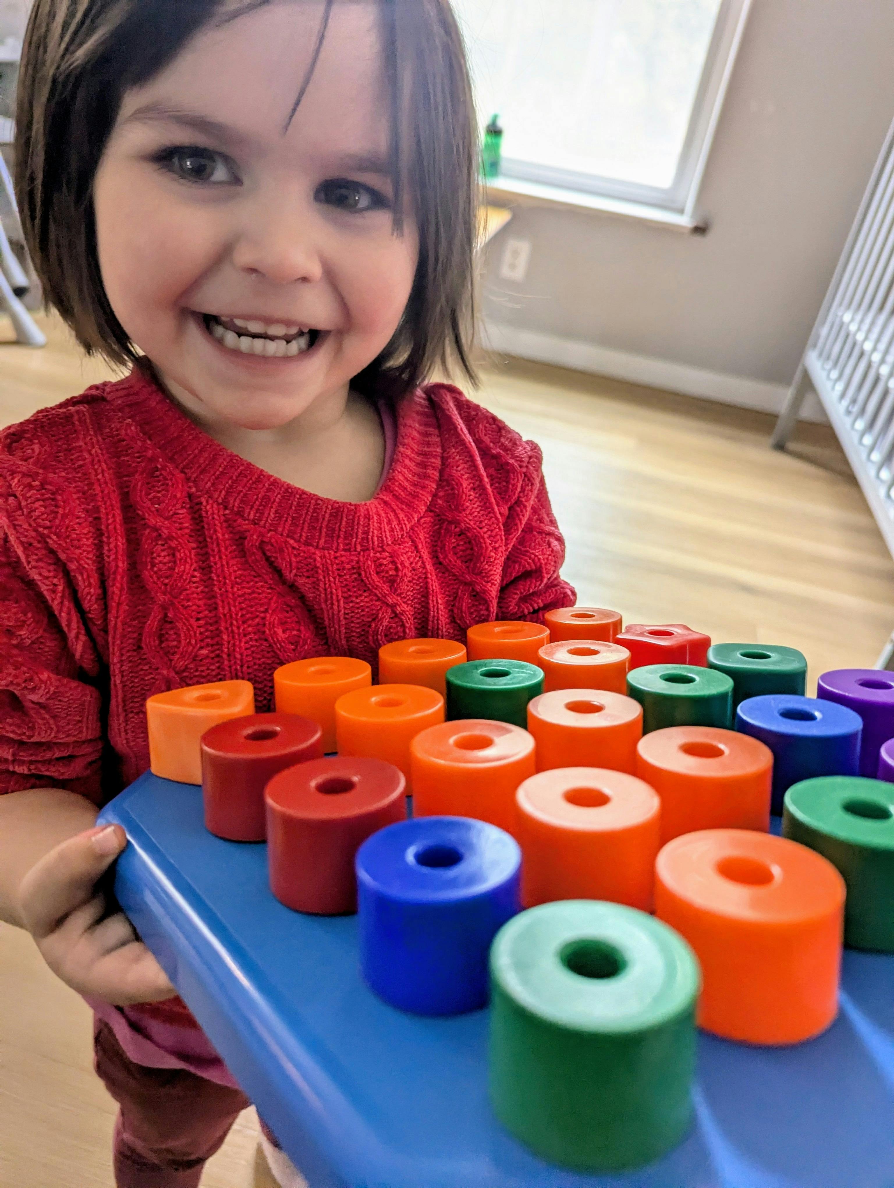 Child in a red sweater holding a blue tray with colorful sorting toys