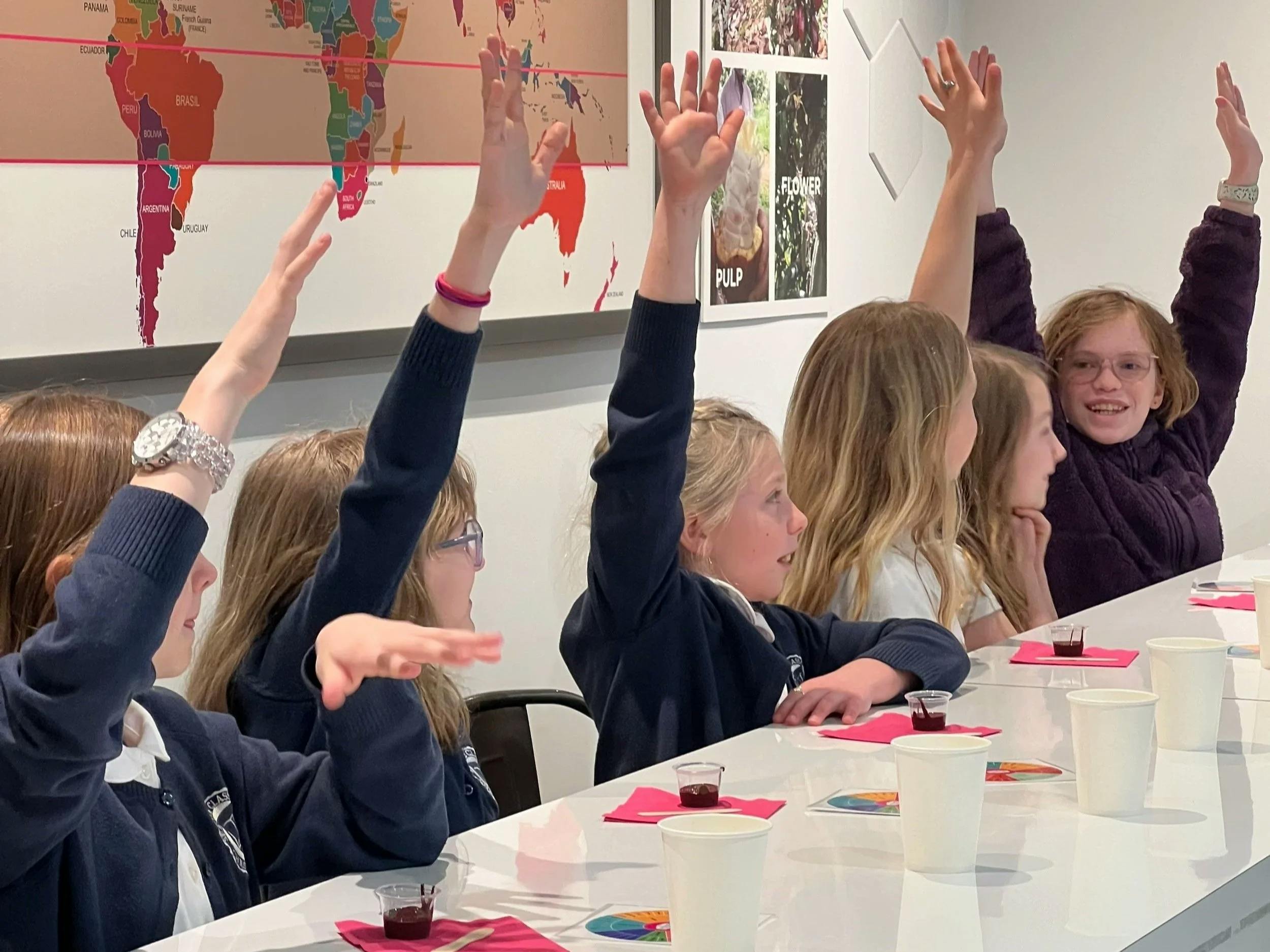 Children raising hands at desks in a classroom with a map behind them