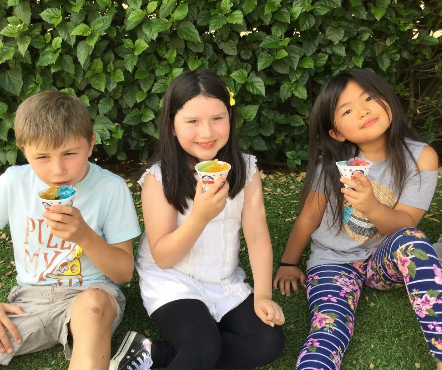Three children sitting on grass holding cups of shaved ice