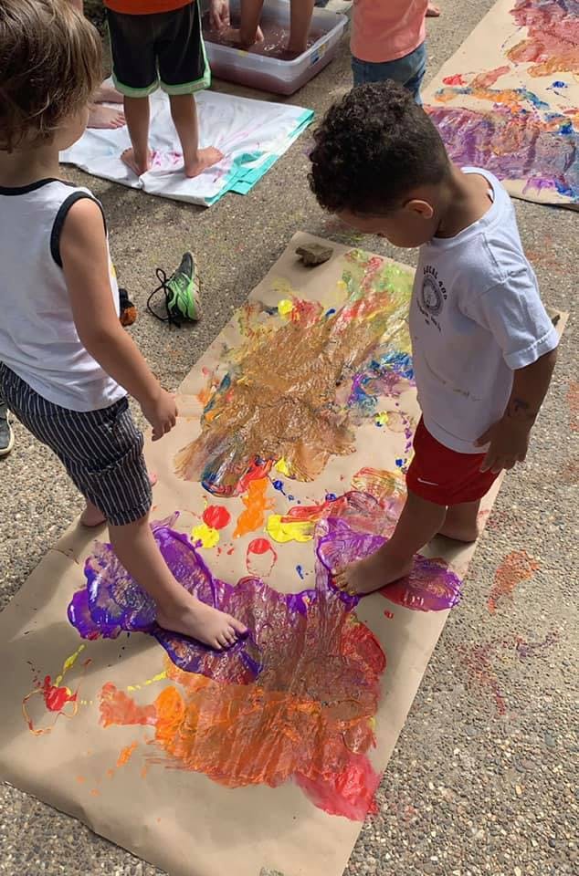 Two children standing barefoot on a large paper with colorful paint