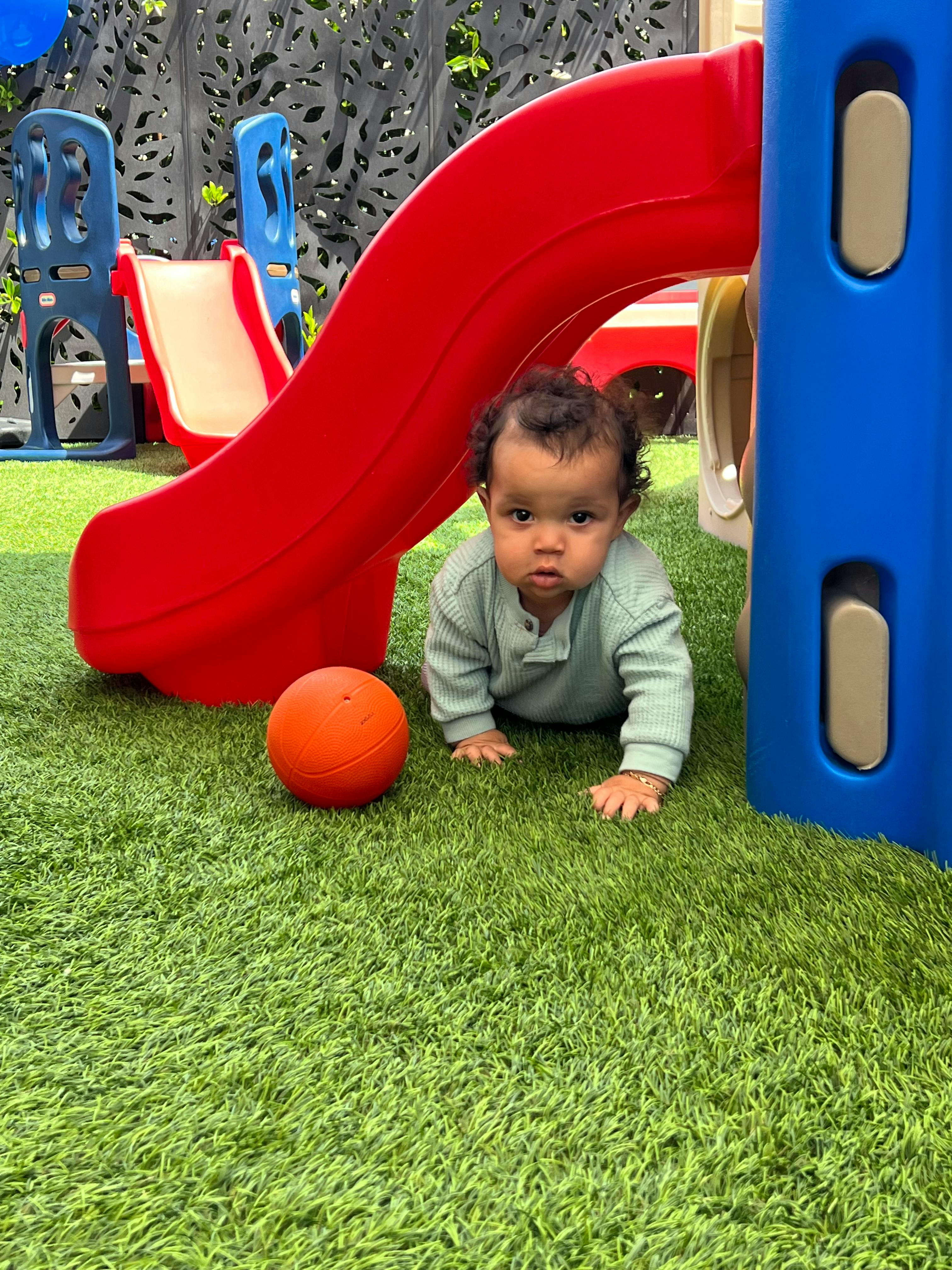 Child crawling on green grass next to an orange ball and playground slide