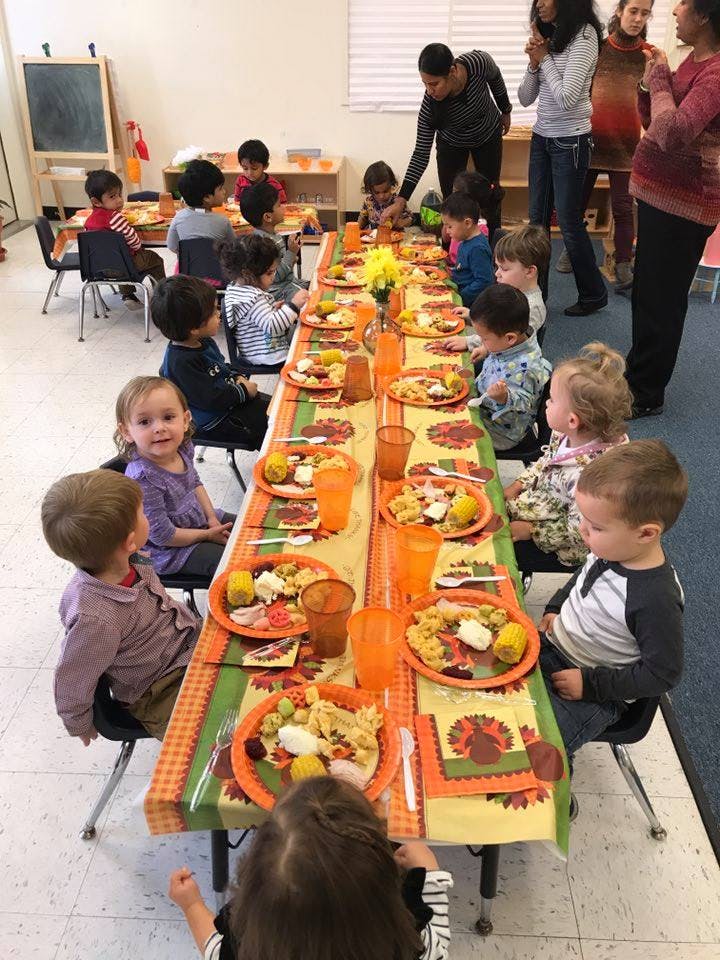 Children seated at a long table with plates of food, adults standing nearby