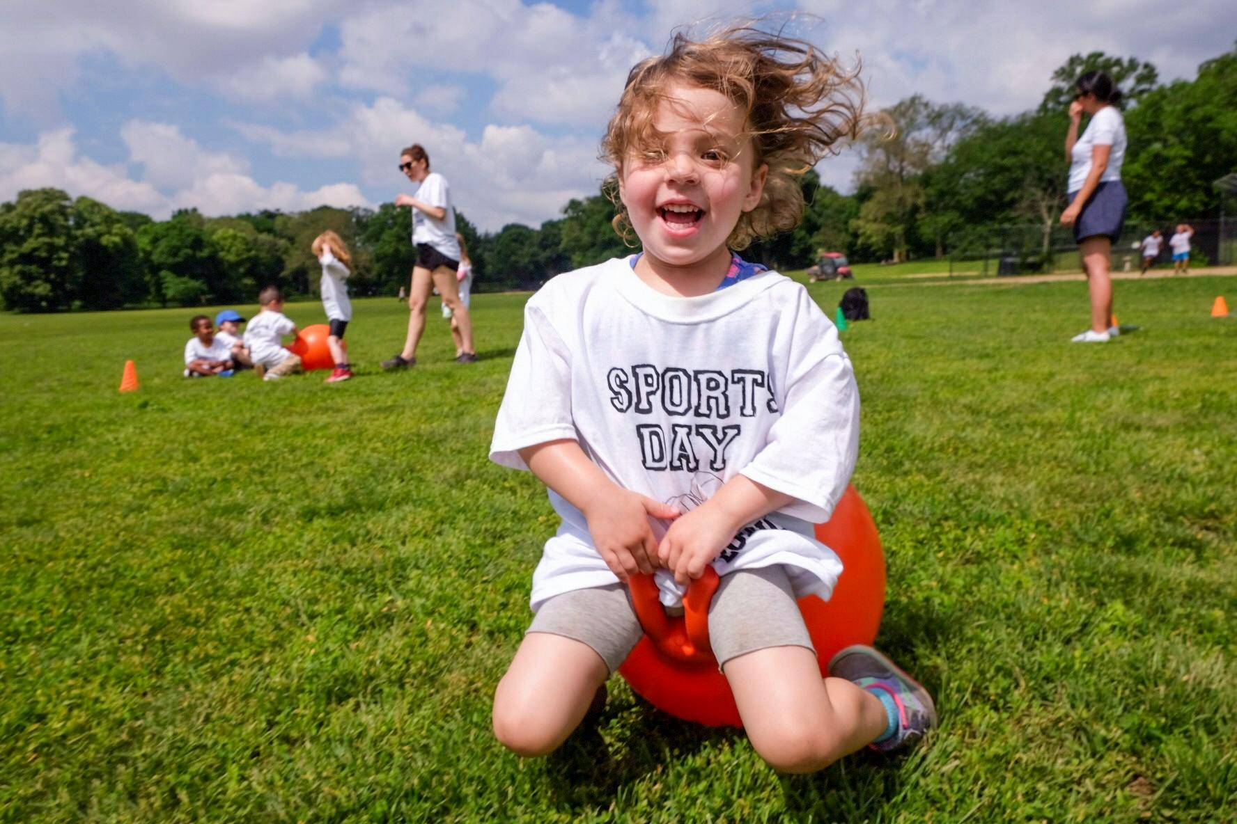 Child in a "Sport Day" shirt kneeling on a red ball in a grassy field