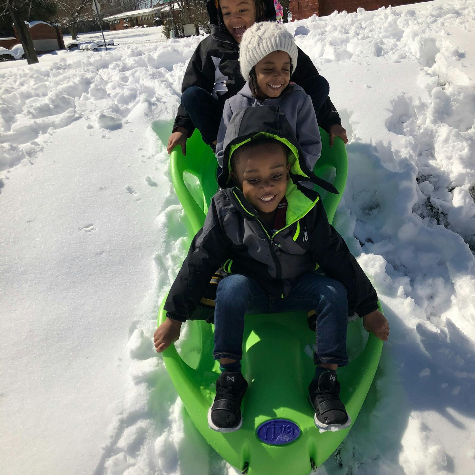 Three children riding together on a green plastic sled in the snow