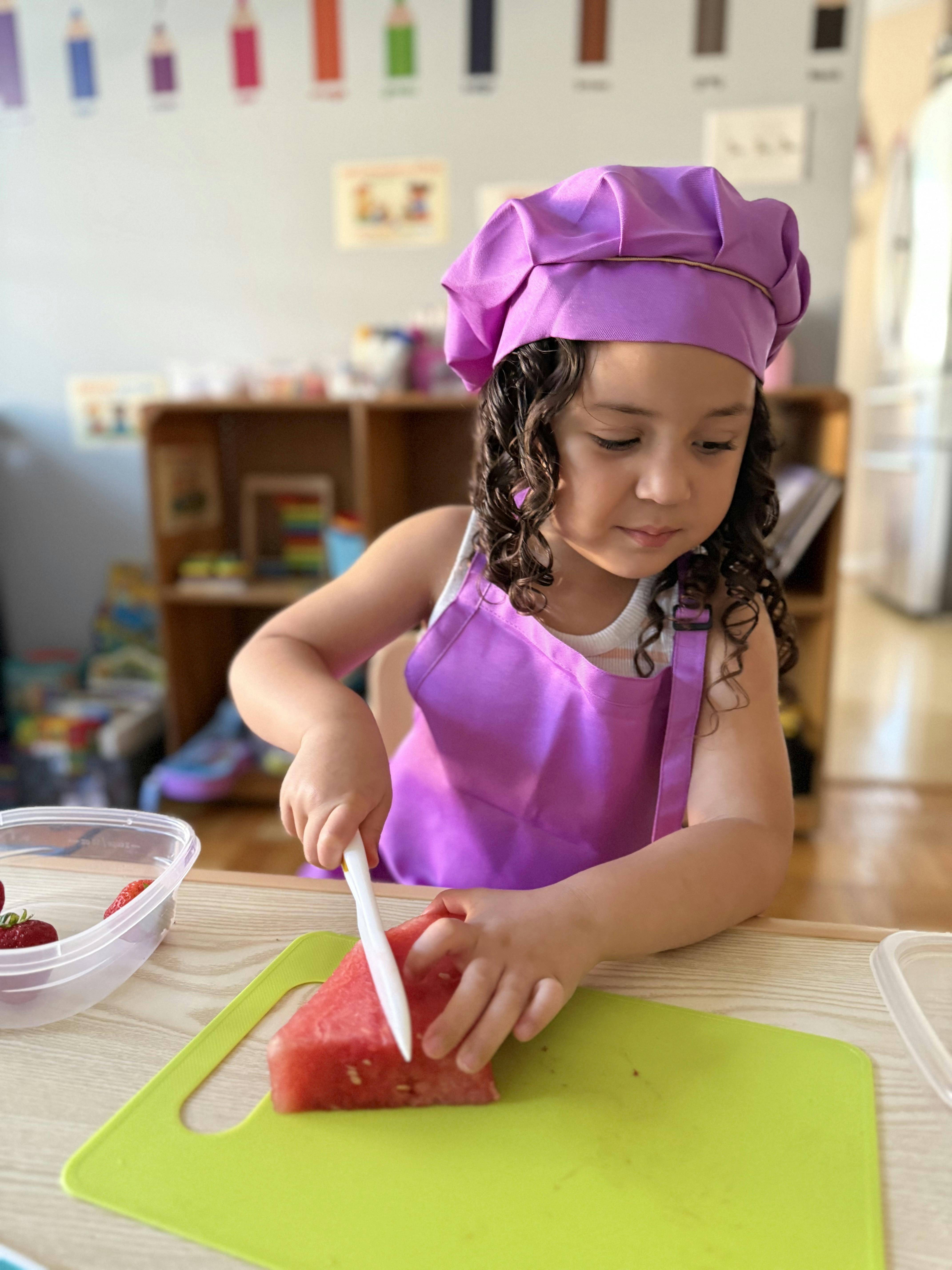 Child in a chef hat and apron cutting watermelon on a cutting board