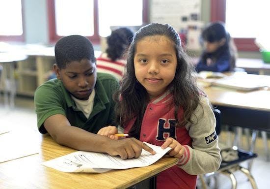 Two children at a desk pointing at a paper in a classroom