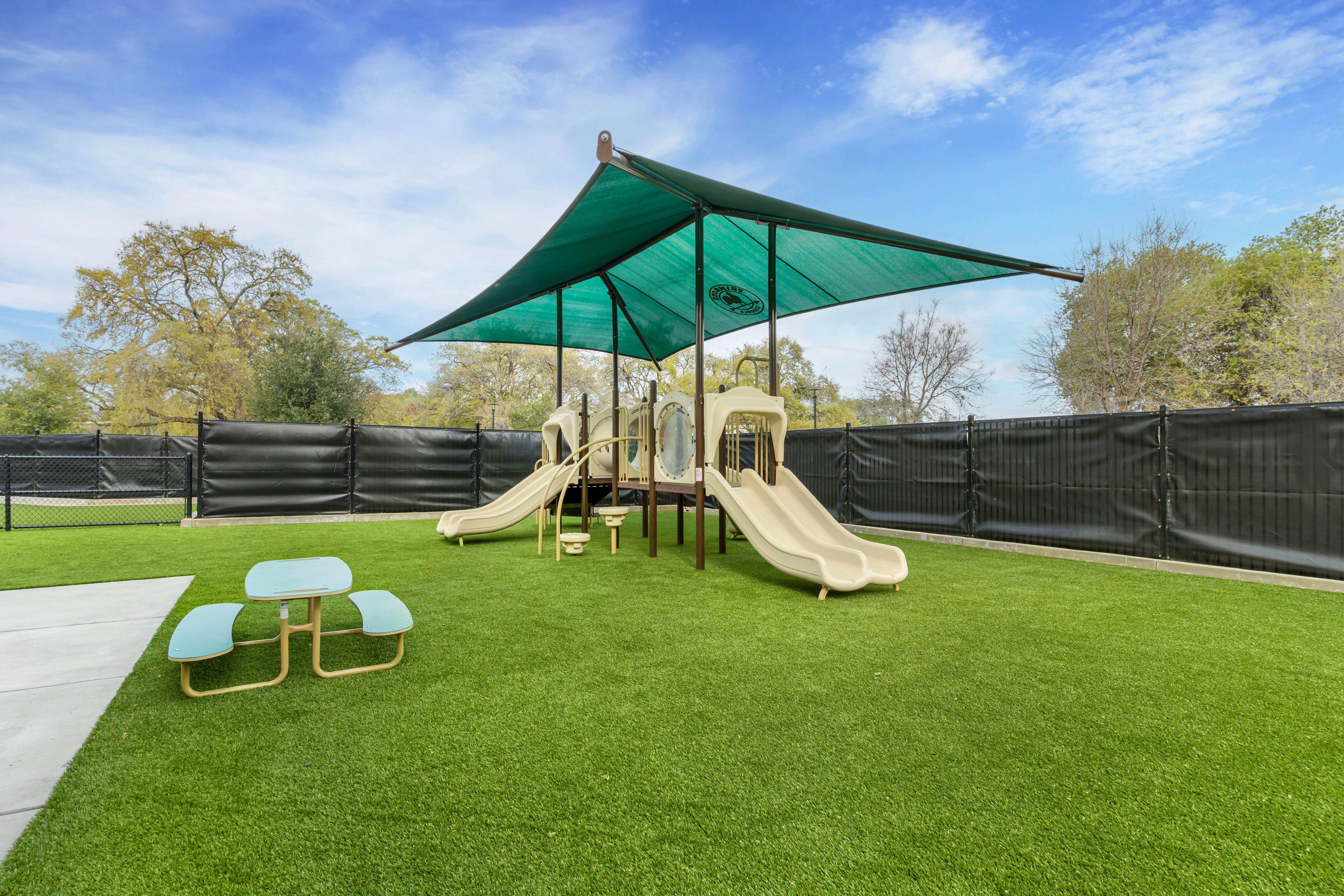 Playground with slides under a green canopy, artificial turf, and a small table