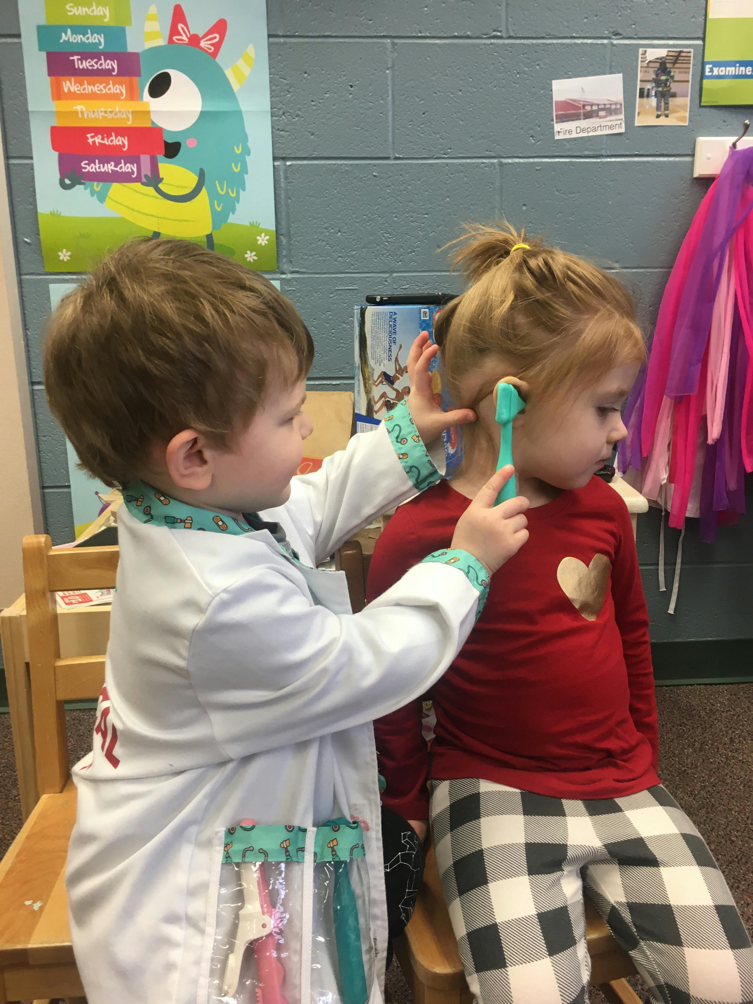 Child in doctor costume examining another child's ear with a toy otoscope