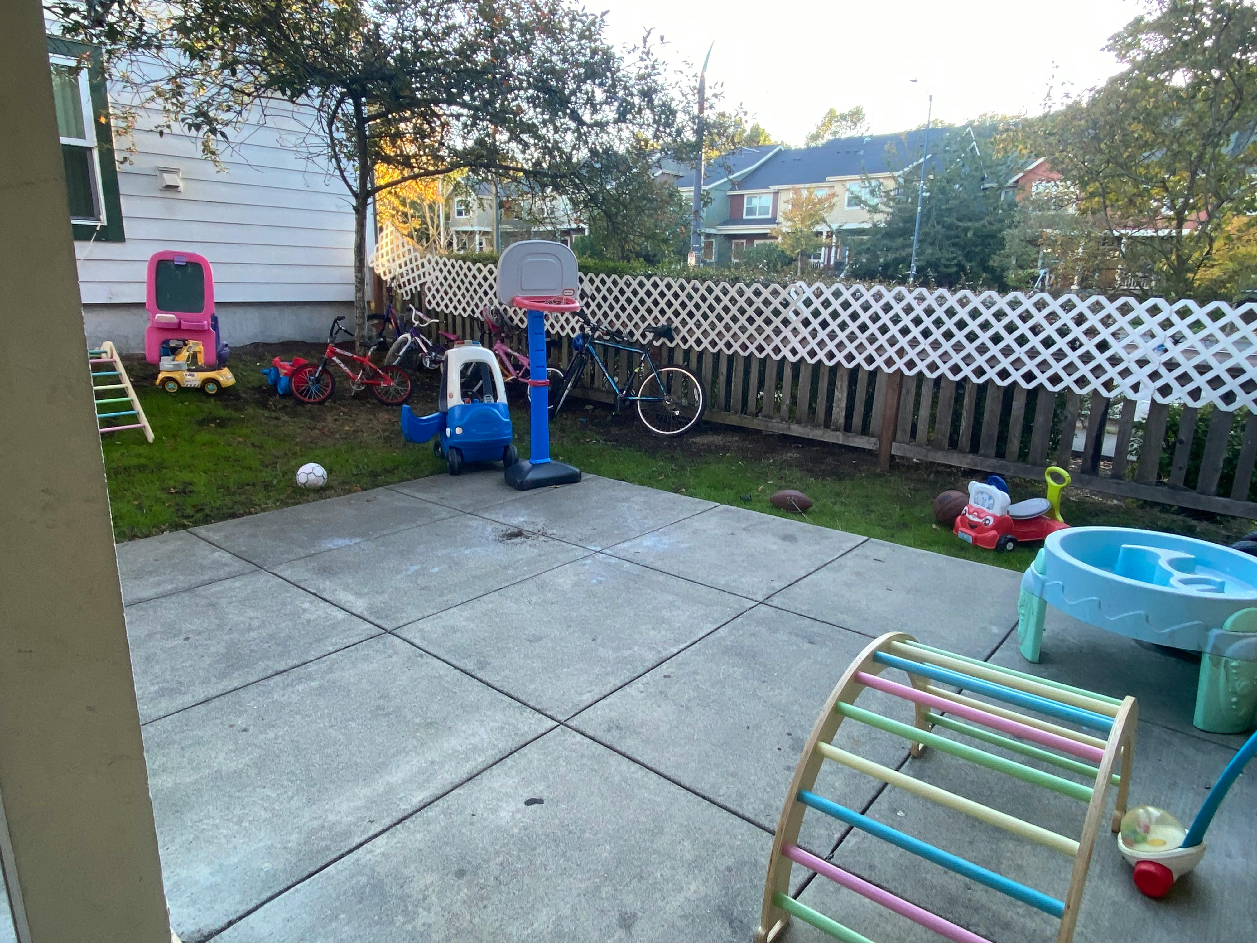 Backyard with a concrete patio, various children's toys, and a white fence