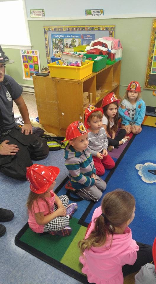 Adult and children in red firefighter hats sitting on a rug in a classroom