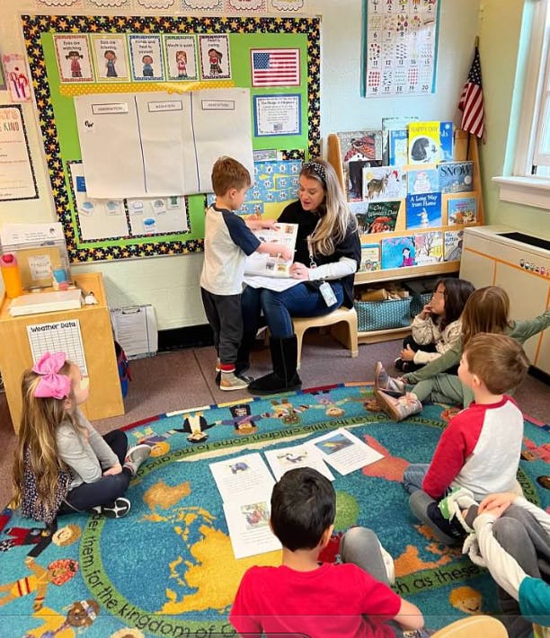 Adult and child looking at a book in a classroom with other children sitting on a rug