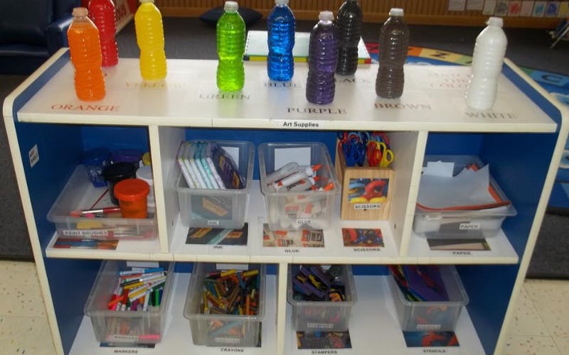 Art supplies shelf with colorful bottles on top, markers, crayons, glue, and paper below