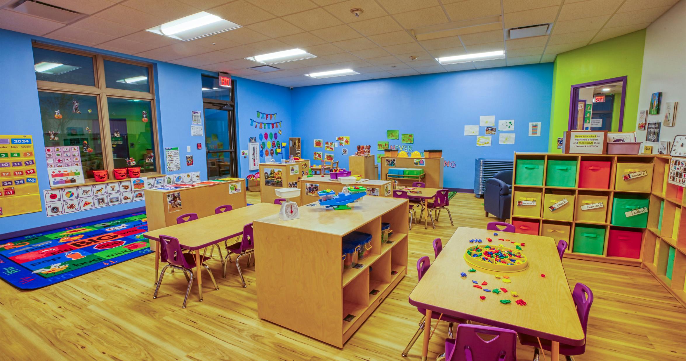 Colorful classroom with small tables and chairs, shelves with bins, and decorations