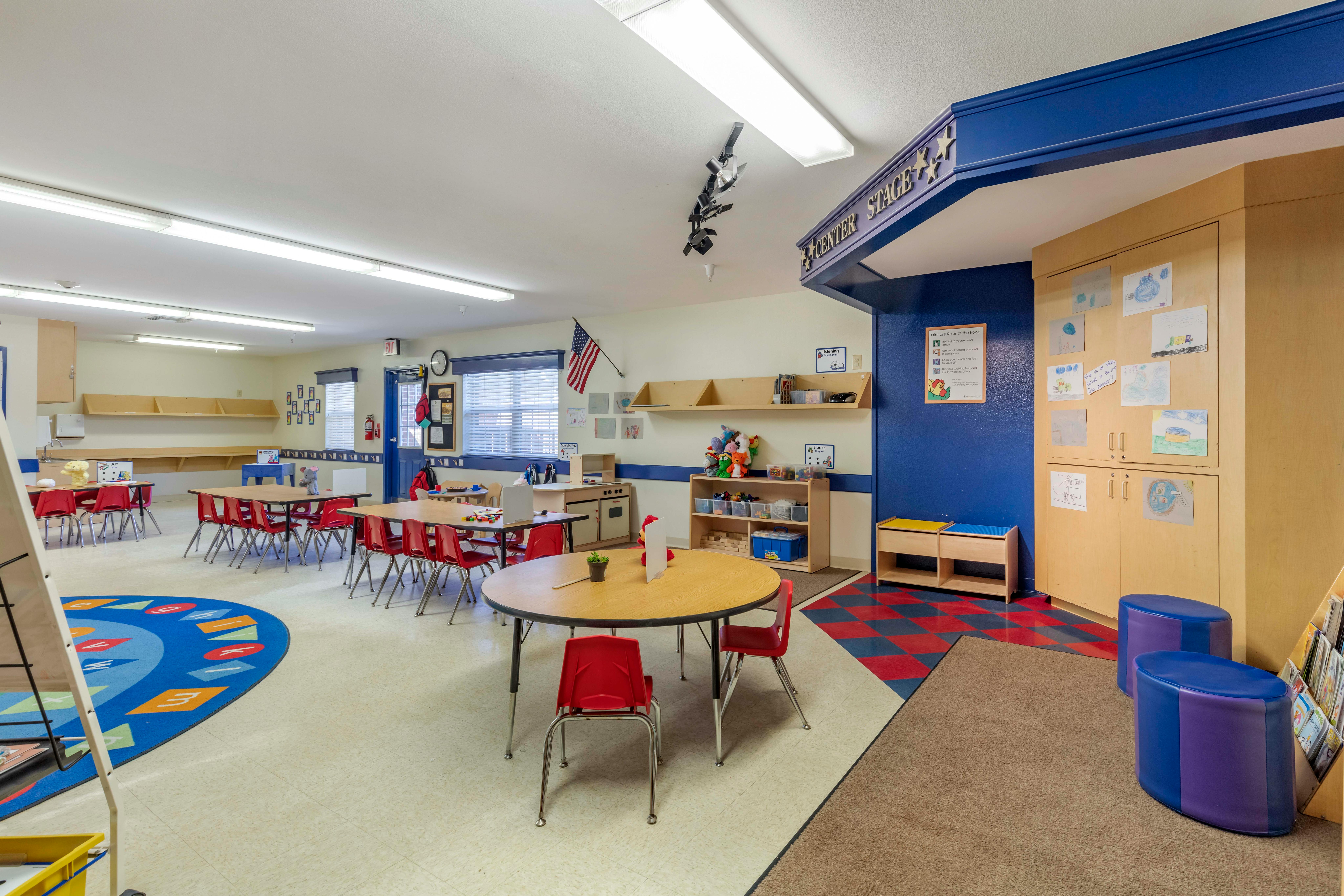 An empty classroom with small tables, red chairs, and a play area