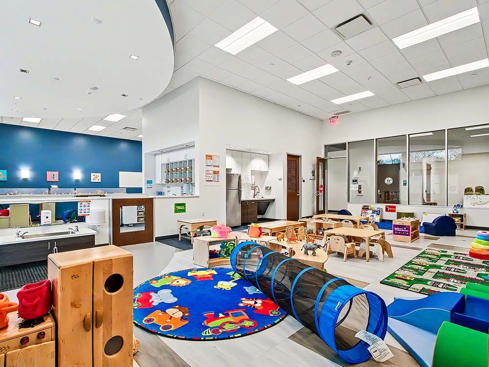 Spacious daycare room with tables, chairs, a play tunnel, and kitchen area