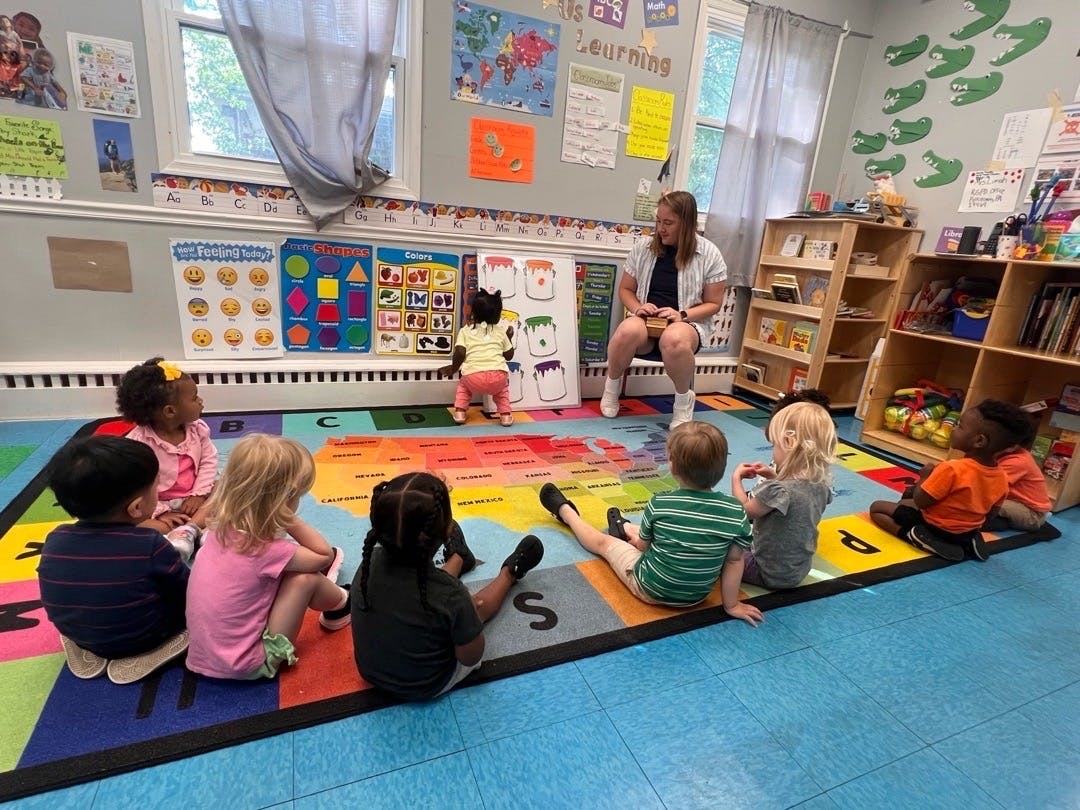 A person leading a group of children sitting on a map rug in a classroom