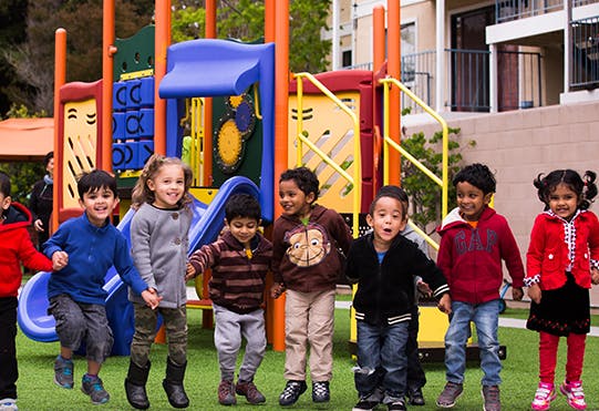 Children holding hands and jumping in front of a playground structure