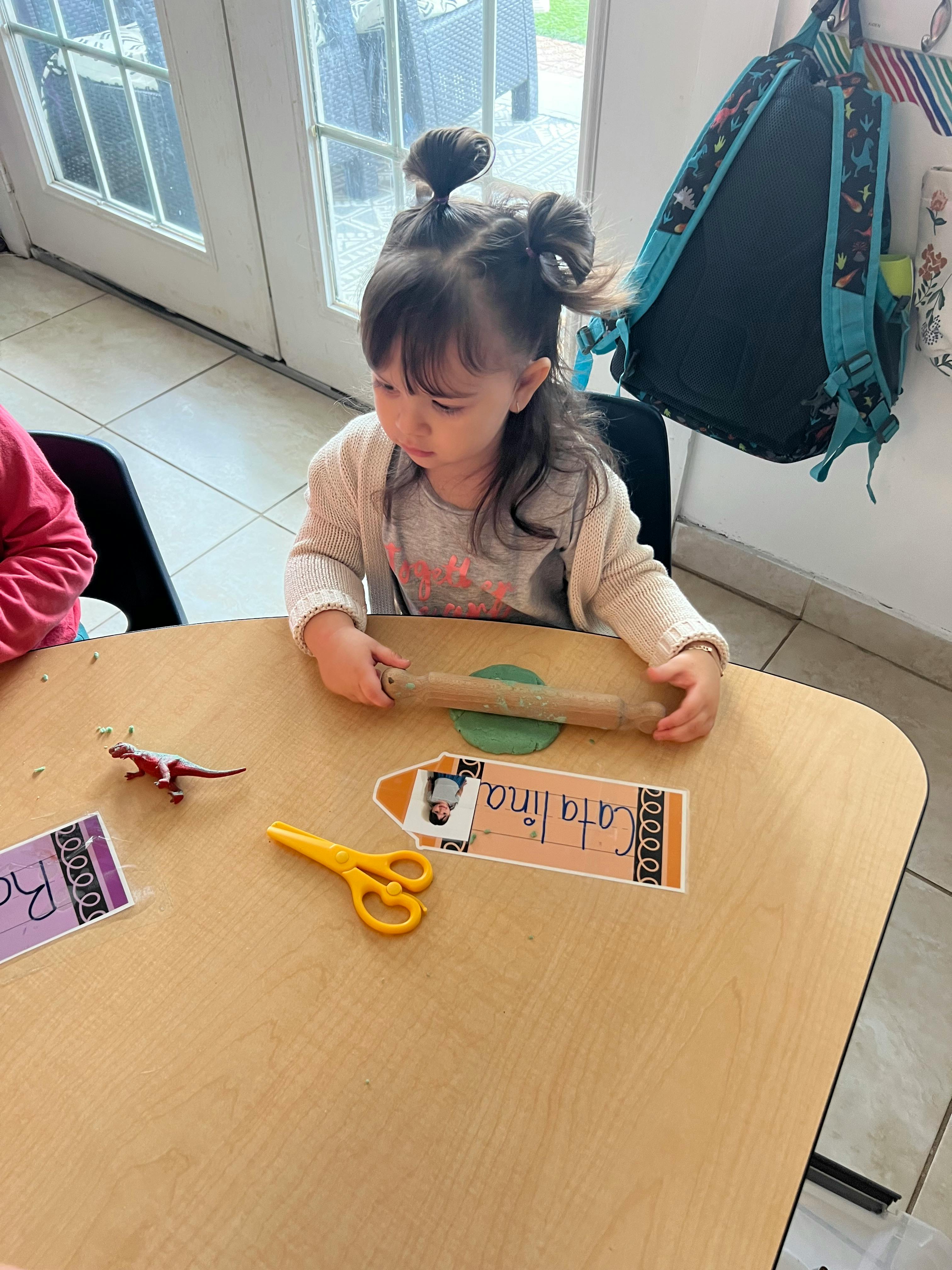 Child using a rolling pin on green playdough at a table