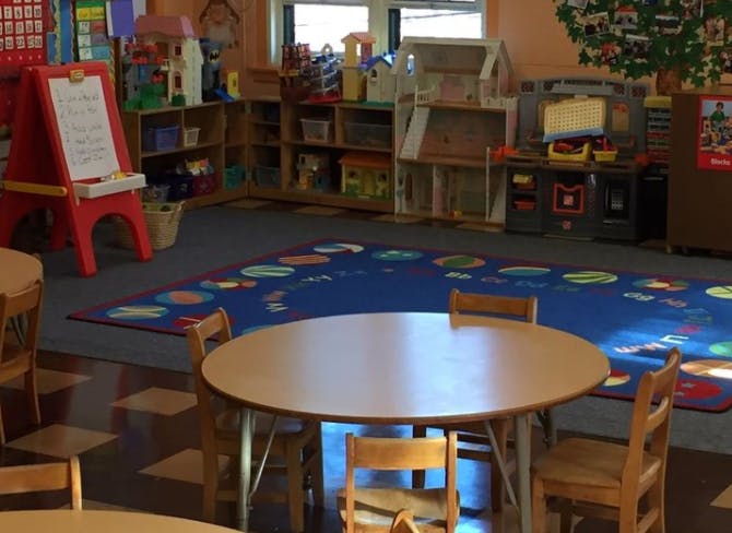 Classroom with small tables and chairs, a blue rug, and shelves with toys