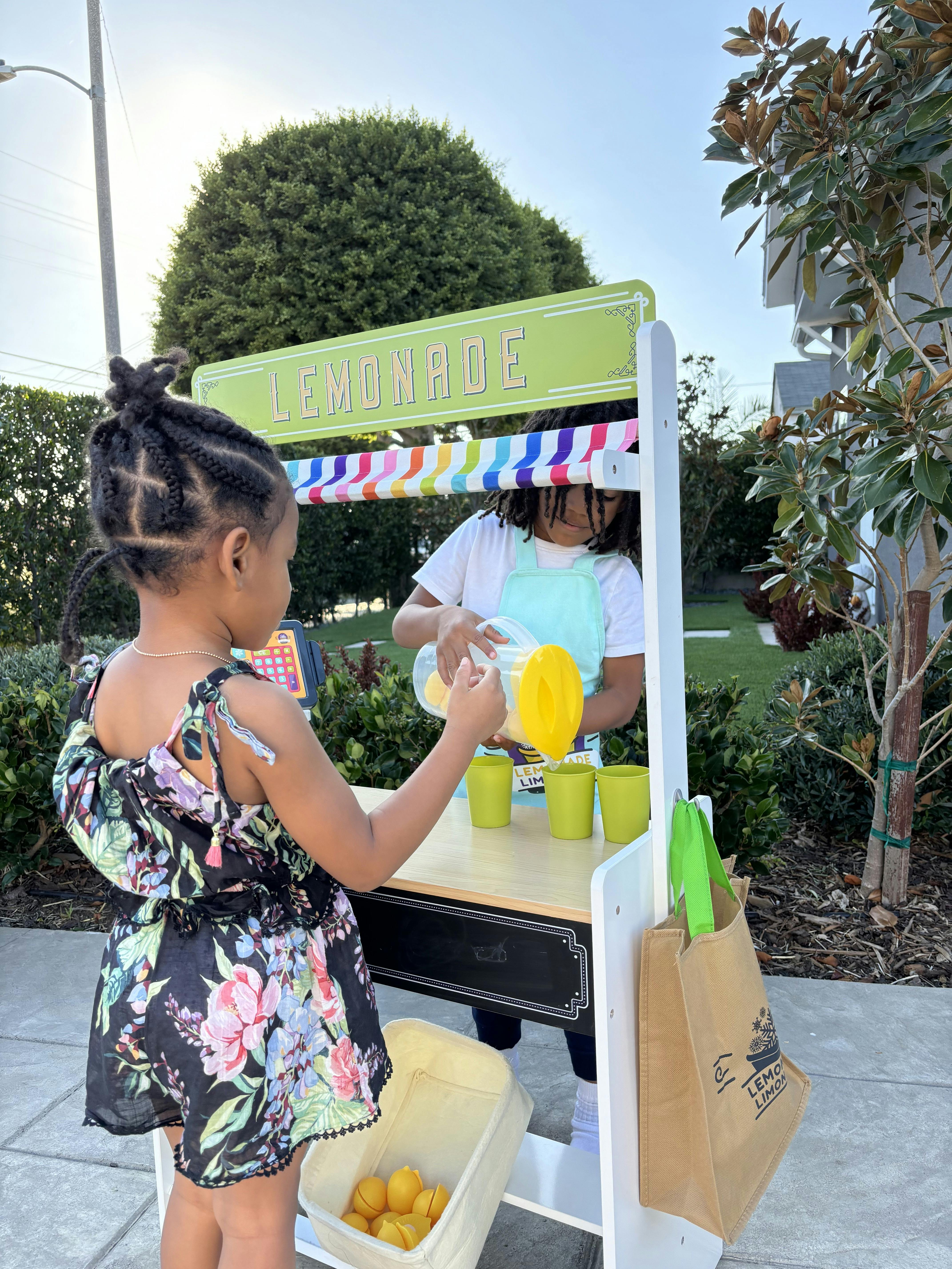 Child pouring from a pitcher at a lemonade stand, with another child watching